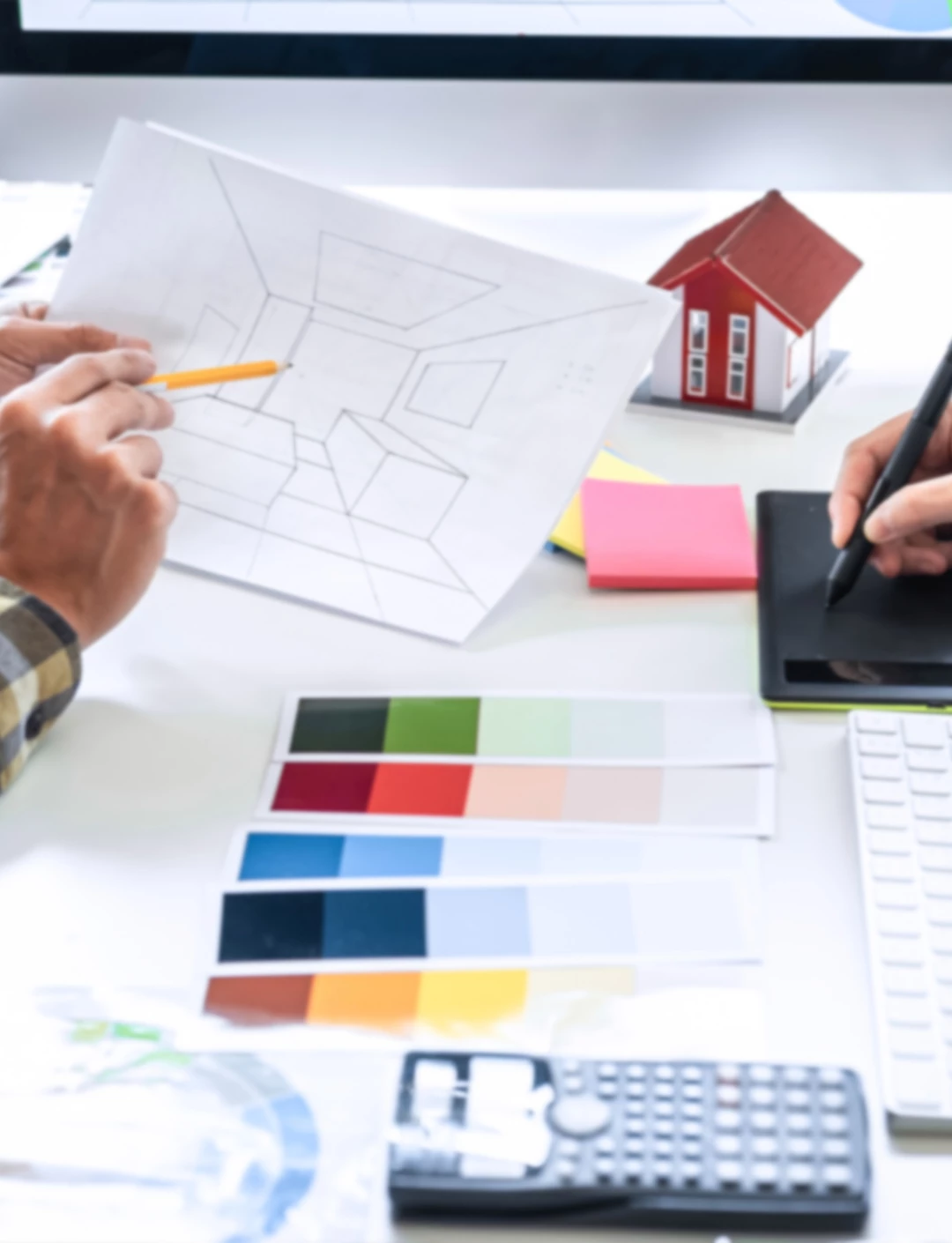 mo segurando um lpis apontando para desenho, paleta de cores em cima da mesa, maquete de casa, calculadora, mesa digitalizadora e papeis adesivos